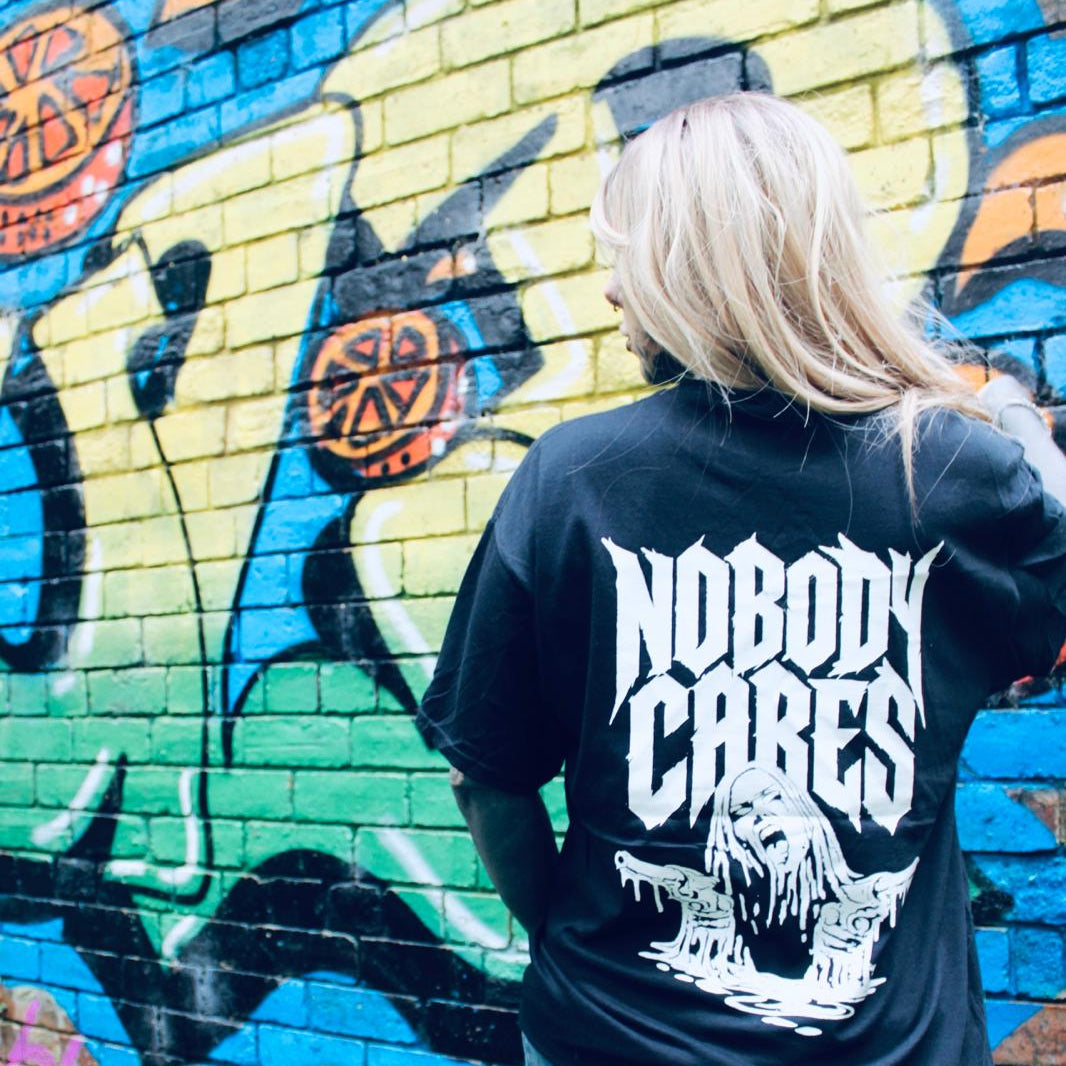 A person with blonde hair seen from behind, wearing a black graphic t-shirt with a large white 'NOBODY CARES' band logo and a skull illustration on the back, standing in front of a colorful graffiti brick wall.