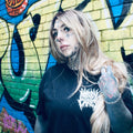 A person with long blonde hair, face tattoos, and a nose ring stands in front of a colorful graffiti brick wall, wearing a black t-shirt with a white graphic logo.
