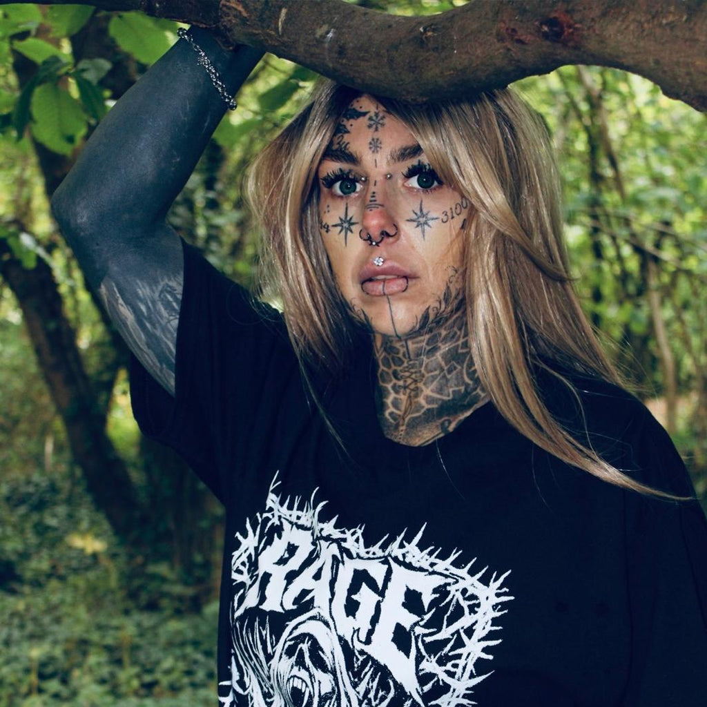 A tattooed woman with face tattoos stands outdoors in a wooded setting, wearing an oversized black graphic T-shirt with a large white gothic-style print reading 'SHRAGE' and 'With No God of Glass.'.