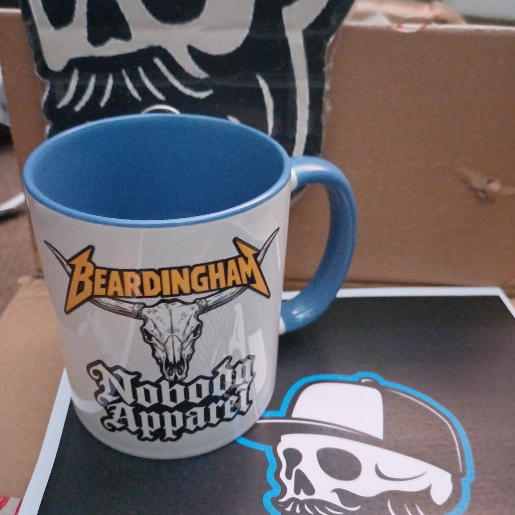 A white ceramic mug with a blue handle and interior, featuring a graphic design with the text 'Beardingham' and 'Wooden Apparel' alongside a skull illustration. The mug is shown resting on a cardboard box next to a matching printed graphic.