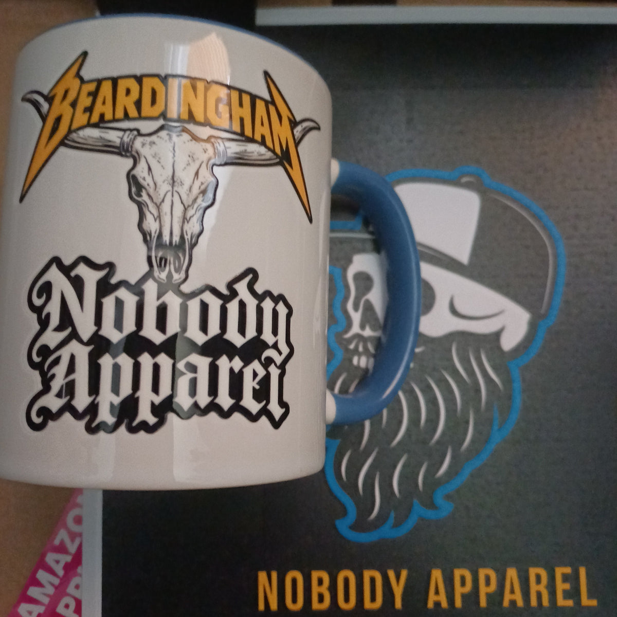 A white ceramic mug with a blue handle, featuring a graphic of a skull with a beard and the text 'Beardingham' and 'Nobody Apparel'. The mug is shown in front of a black box with a matching graphic and the words 'NOBODY APPAREL'.