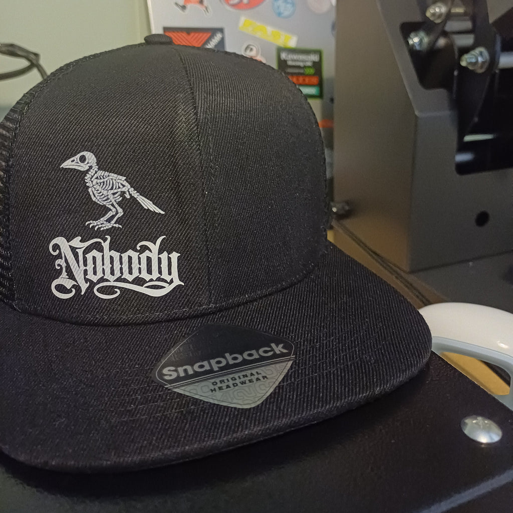 A black trucker-style cap with a white embroidered skeleton bird and gothic-style text on the front, photographed indoors near a sticker-covered surface and a dark machine component.