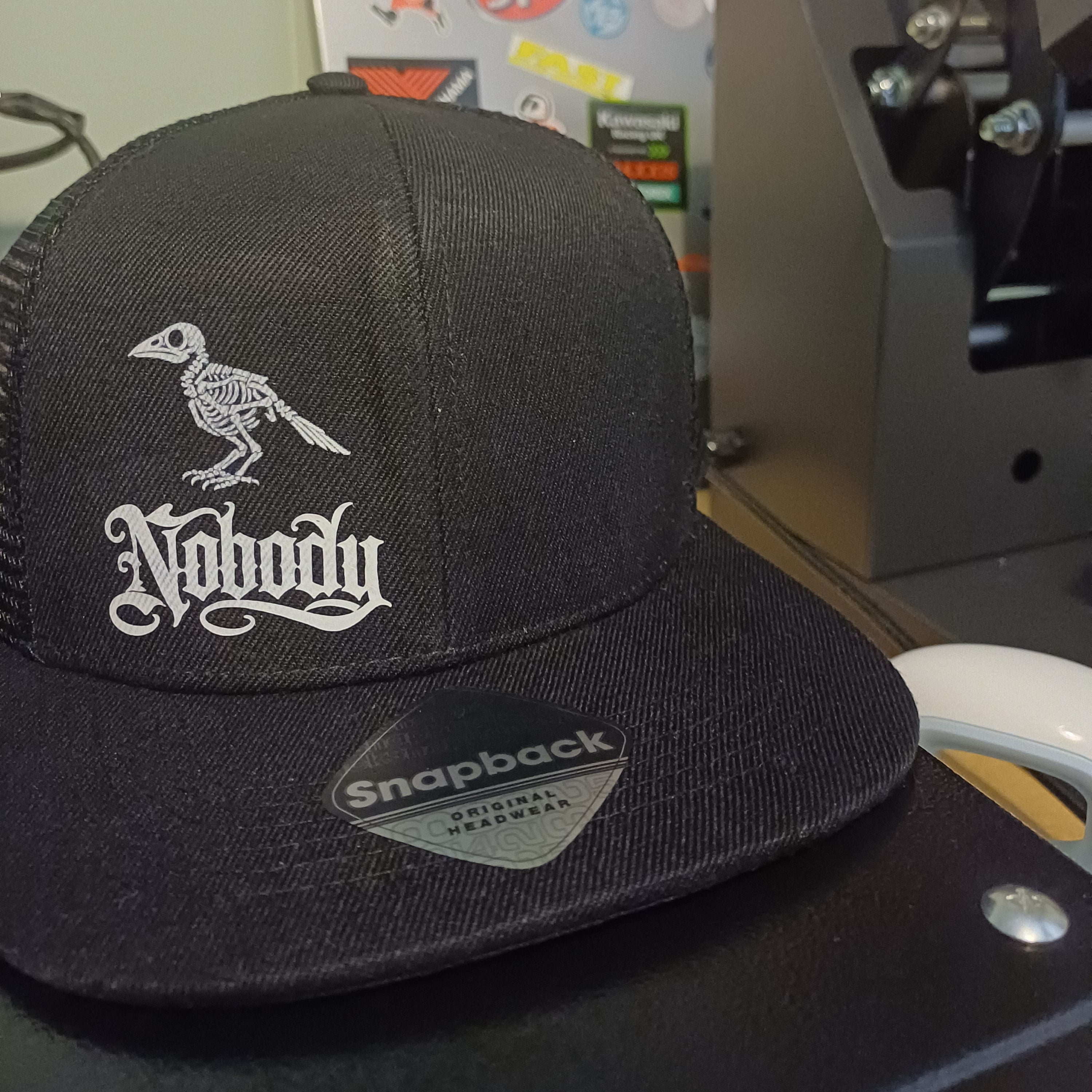 A black trucker-style cap with a white embroidered skeleton bird and gothic-style text on the front, photographed indoors near a sticker-covered surface and a dark machine component.
