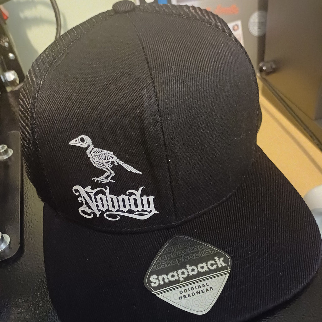 A black trucker-style cap with a white embroidered bird logo and gothic text on the front panel, plus a silver and black Snapback sticker on the brim, photographed indoors on a dark surface.