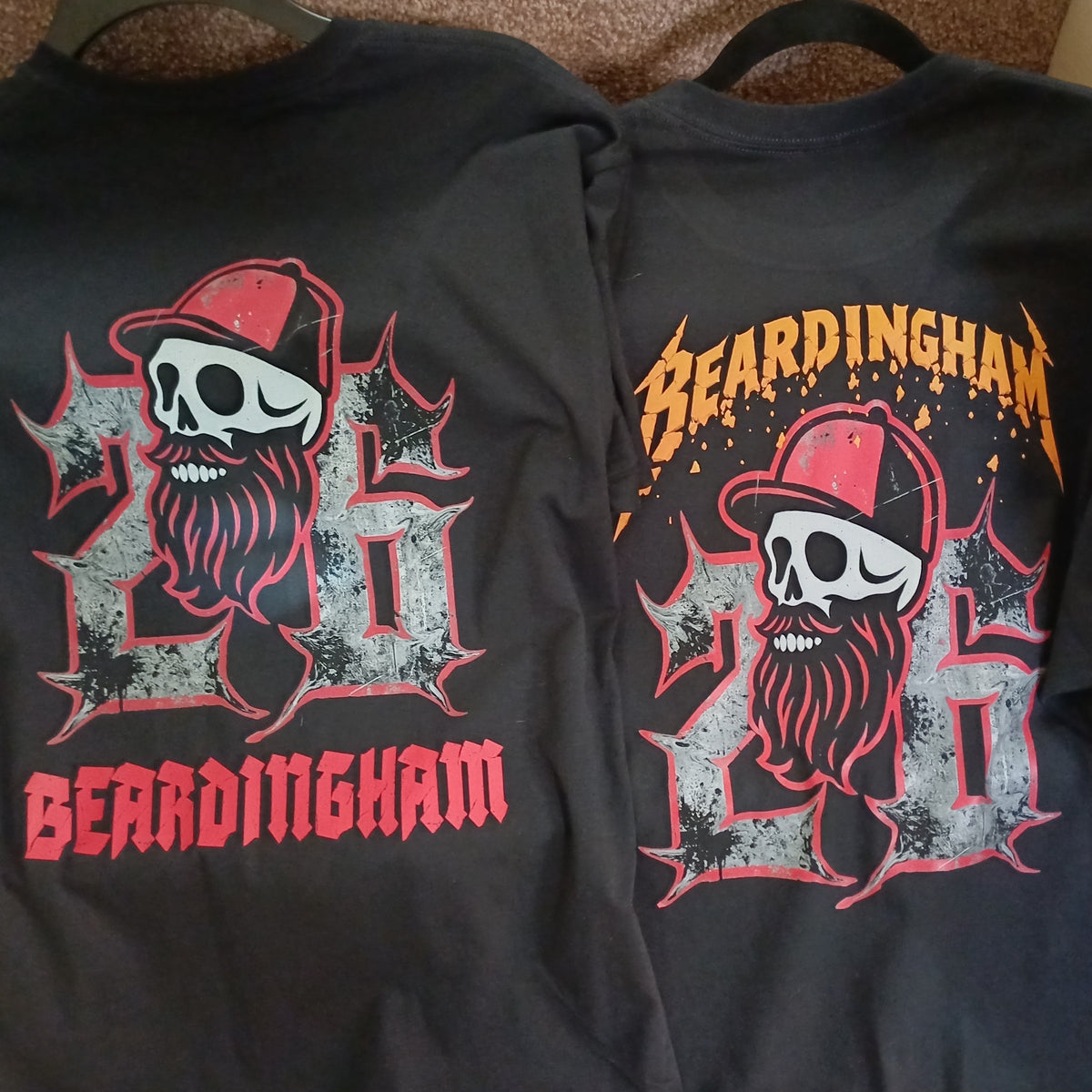 Two black short-sleeve t-shirts laid flat side by side, showing the front and back graphics. The shirts feature a skull wearing a red cap, a large distressed gray '26', a red beard, and 'BEARDINGHAM' text in red and orange.