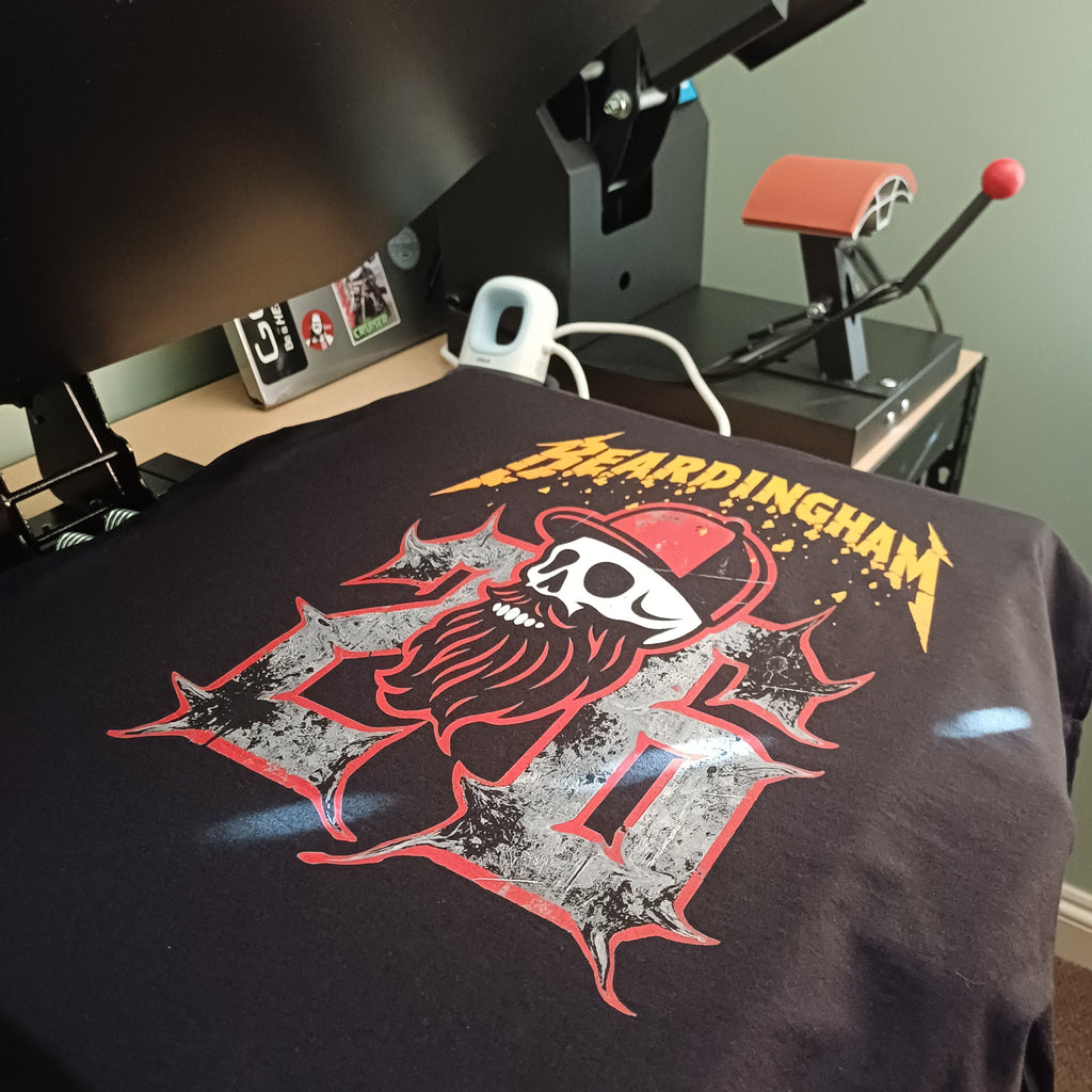A black t-shirt with a 'Bardingham III' graphic print is being held under a heat press machine in a workshop setting. The graphic features a skull with a red bandana and a large distressed 'III' in red and gray, with 'BARDINGHAM' in yellow text above.