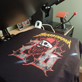 A black t-shirt with a 'Bardingham III' graphic print is being held under a heat press machine in a workshop setting. The graphic features a skull with a red bandana and a large distressed 'III' in red and gray, with 'BARDINGHAM' in yellow text above.
