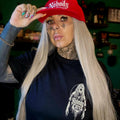 A woman with long platinum blonde hair, face tattoos, and a nose ring wears a red baseball cap with the word 'Nobody' in white script and a dark navy graphic t-shirt. She is posing indoors against a dark green wall.