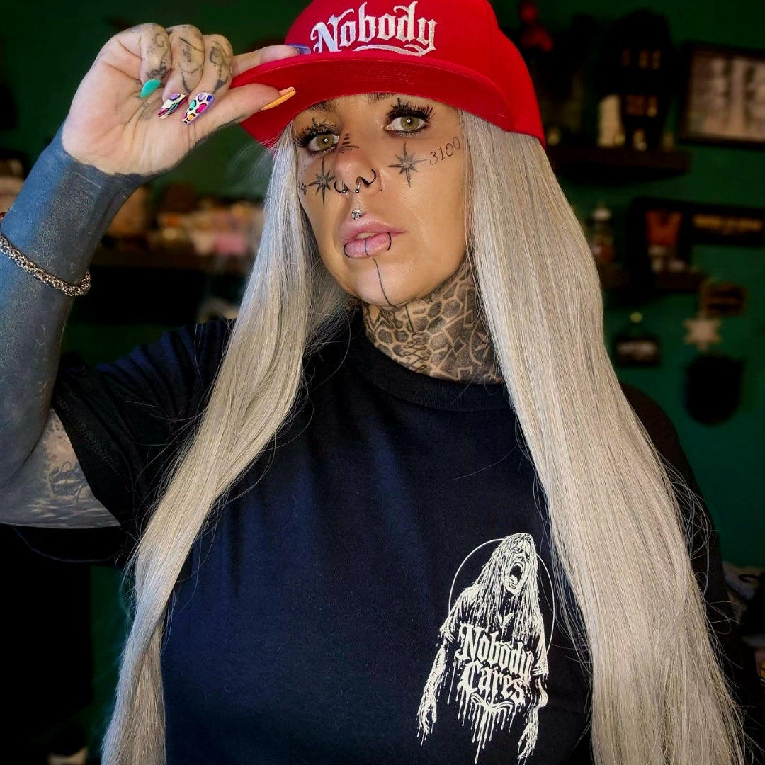 A woman with long platinum blonde hair, face tattoos, and a nose ring wears a red baseball cap with the word 'Nobody' in white script and a dark navy graphic t-shirt. She is posing indoors against a dark green wall.