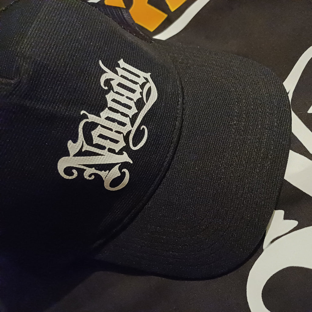 A close-up of a black baseball cap featuring a white Gothic-style embroidered logo that reads 'Nobody' on the front panel, resting on a black garment with a large white gothic graphic.