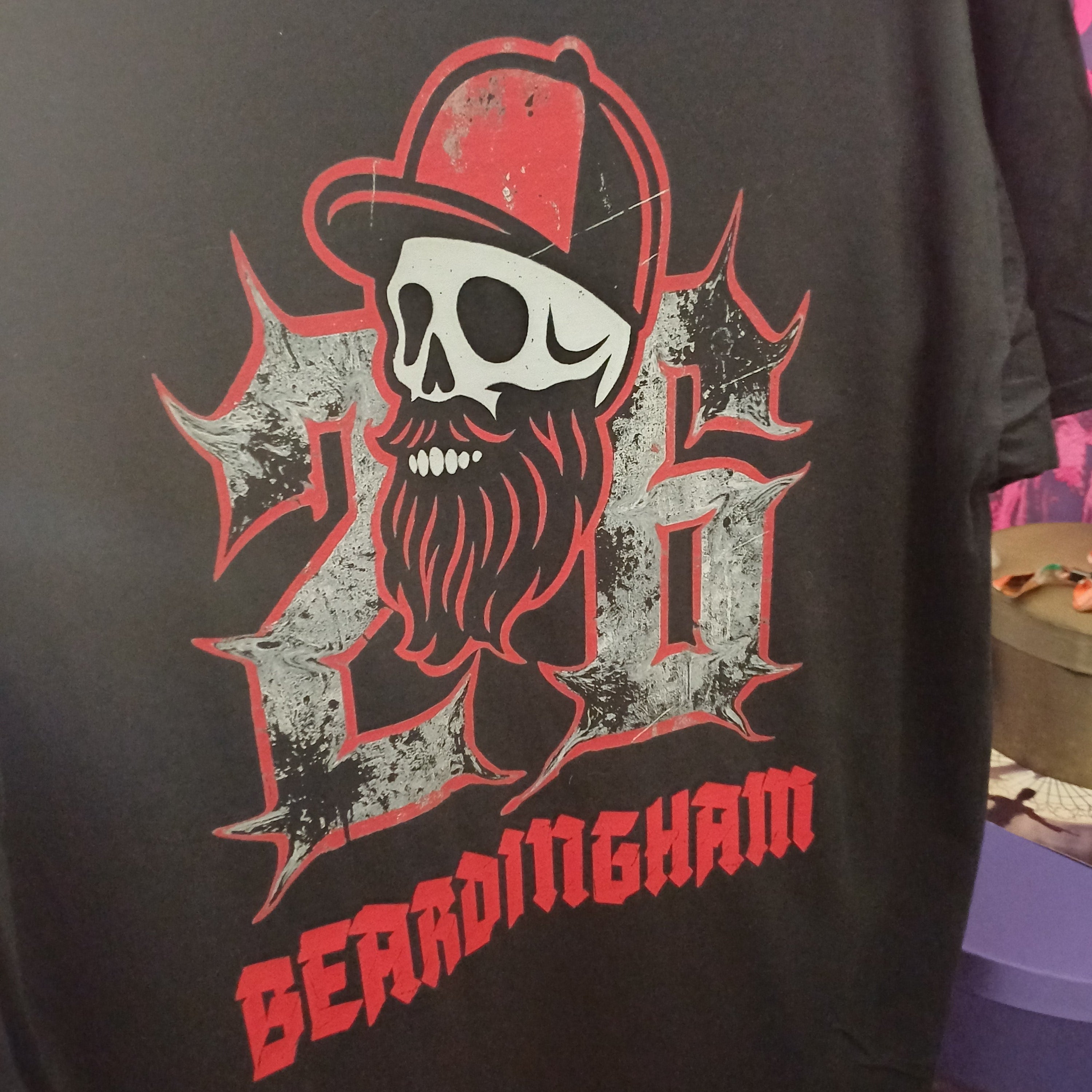 A close-up of a black t-shirt featuring a graphic print of a skull with a red cap and a long beard, integrated into the number 26, with the word 'BEARDINGHAM' printed in red below.