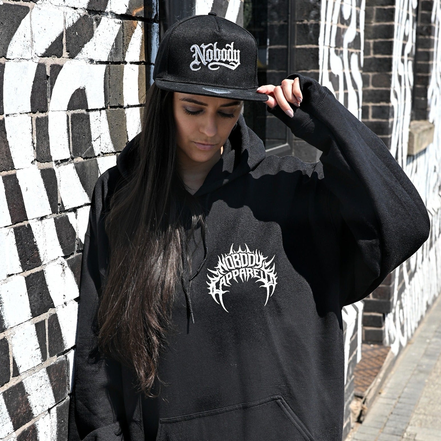 A woman wearing a black hoodie with a white graphic logo and a matching black cap with 'Nobody' in white gothic lettering, standing against a black and white patterned brick wall.