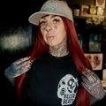 Person with tattoos wearing a cap and black t-shirt with a logo in a room with posters on the wall.