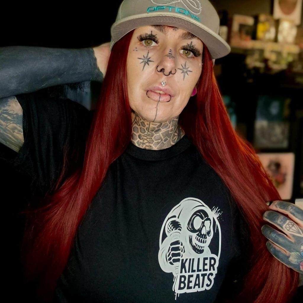 Person with red hair and tattoos wearing a cap and 'Killer Beats' t-shirt in a tattoo parlor.