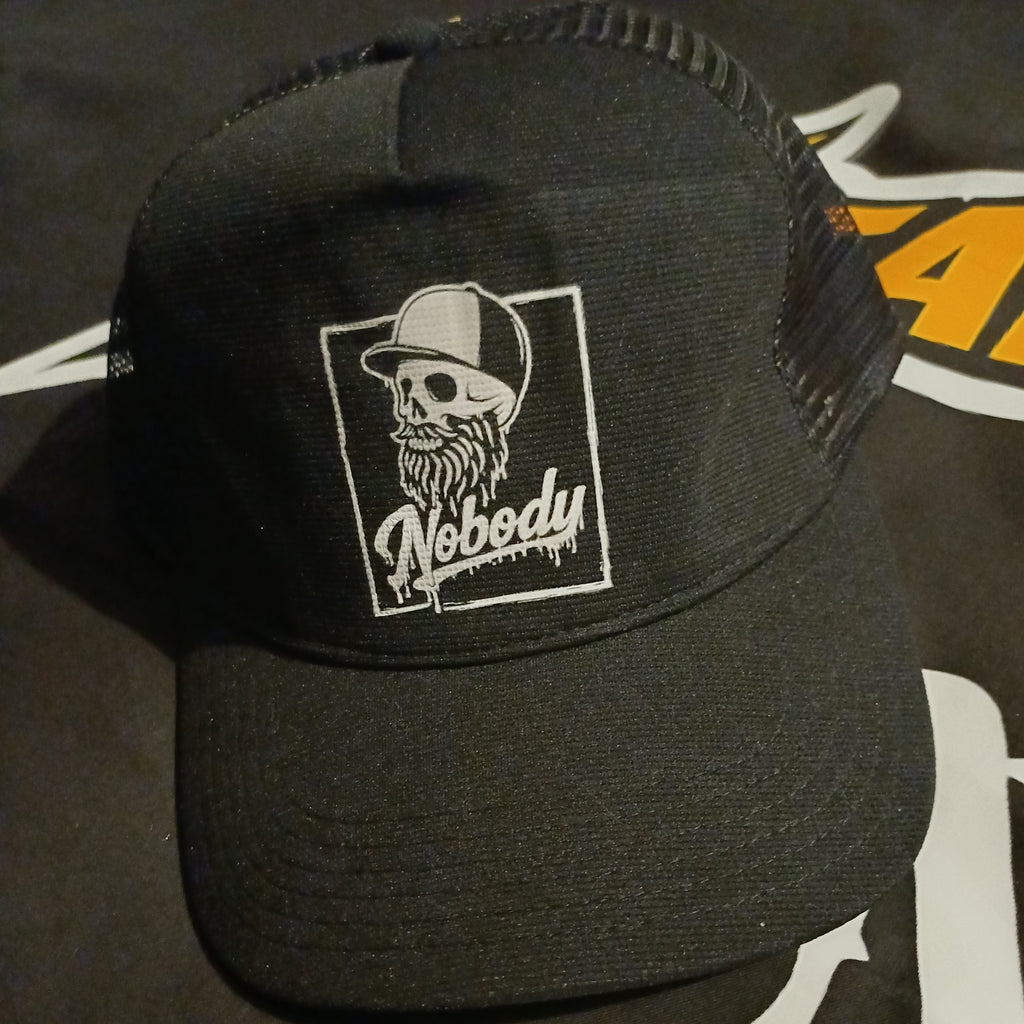 A black trucker hat with a white embroidered patch on the front featuring a skull wearing a cap and the word 'Nobody'.