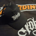 A black trucker-style baseball cap with white gothic embroidery reading 'Naginata' on the front, displayed on a black fabric backdrop with orange and white graphic lettering.