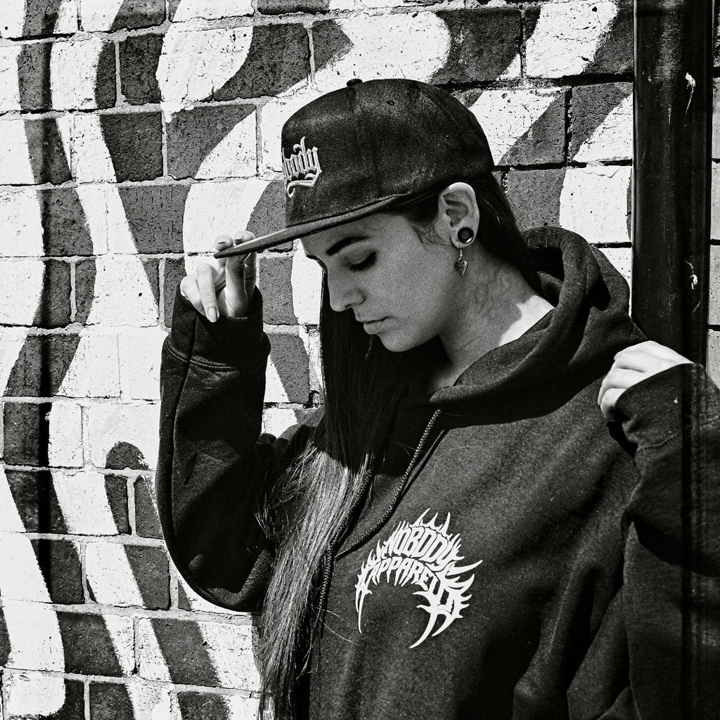 A person in a black hoodie and baseball cap stands against a graffiti-covered brick wall, looking downward. The hoodie has a white graphic on the chest, and the photo is in black and white.