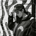 A person in a black hoodie and baseball cap stands against a graffiti-covered brick wall, looking downward. The hoodie has a white graphic on the chest, and the photo is in black and white.