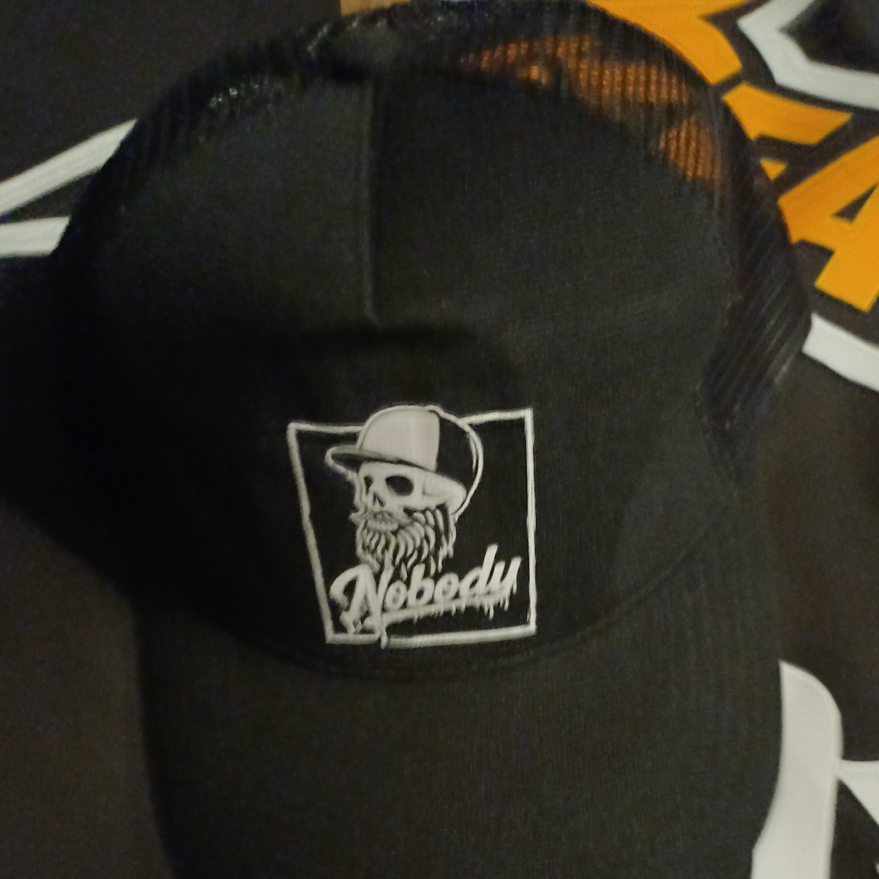 A black trucker hat with a white embroidered patch featuring a skull wearing a baseball cap and the word 'Nobody' on the front, set against a dark background with a graphic print.