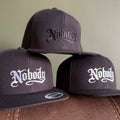 Three dark charcoal snapback caps with 'Nobody' embroidered in Gothic lettering, displayed on a brown leather surface against a beige wall.